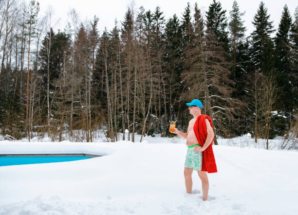 a middle-aged man stands in swimming trunks and with a cocktail in his hand against the background of a winter landscape. - tourist resort fotos imagens e fotografias de stock