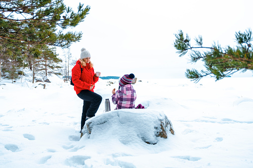 Young woman and child are resting on a picnic in winter. A Caucasian family is drinking hot tea with sandwiches. Picnic on the shores of the frozen bay.
