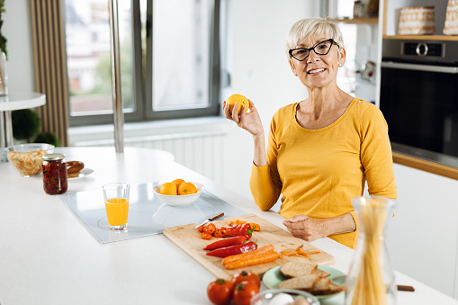 Happy senior woman holding orange in the kitchen and looking at camera