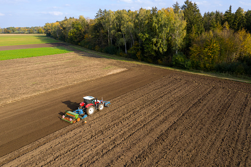 A red, modern, agricultural tractor cultivating and sowing field in a beautiful, idyllic landscape, aerial view.