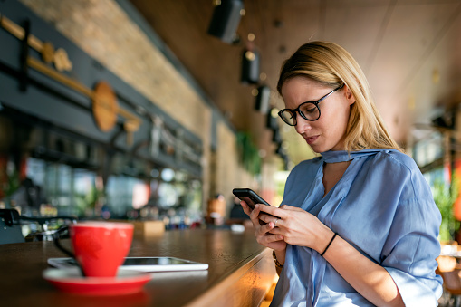 A Serious Millennial Business Woman with Eyeglasses has a Coffee Break in a Bar and Typing Text Messages on her Phone. A Modern and Fashionable Blonde Woman is Using a Smart Phone and Drinking Coffee in Modern Restaurant.