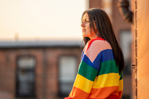 A woman wearing a rainbow sweater stands next to a brick building and poses for a portrait confidently.