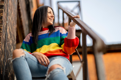A woman wearing a rainbow sweater stands next to a brick building and poses for a portrait confidently.