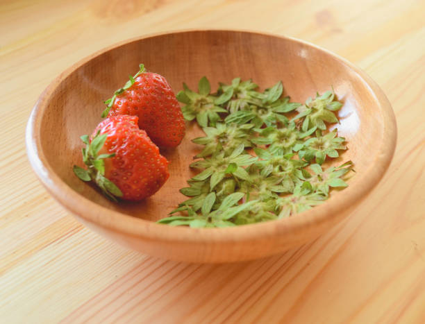 Half eaten strawberry and whole strawberry with calyx in a wooden bowl on a wooden table stock photo