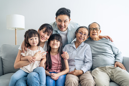 Portrait of Asian happy family sit on sofa and smile, look at camera. Young couple parents spend time with little daughter sibling and senior elder grandparent in house. Multi-generation relationship.