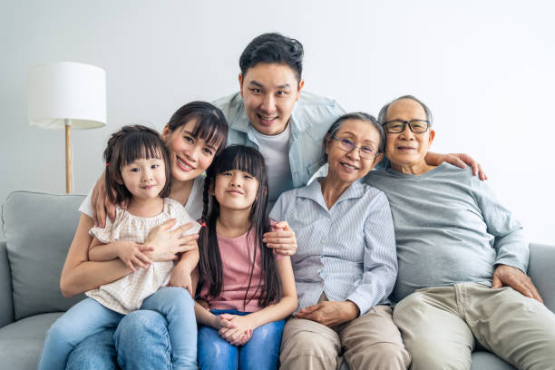portret azjatyckiej szczęśliwej rodziny siedzieć na kanapie i uśmiechać się, spojrzeć na kamerę. młodzi rodzice pary spędzają czas z rodzeństwem córeczki i starszym dziadkiem w domu. relacja wielopokoleniowa. - multi family zdjęcia i obrazy z banku zdjęć