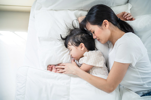 Asian happy family stay home, mother sleep with daughter in bedroom. Attractive loving parent, mom spend time and hug sleeping young little girl child baby with gentle on bed in the morning in house.