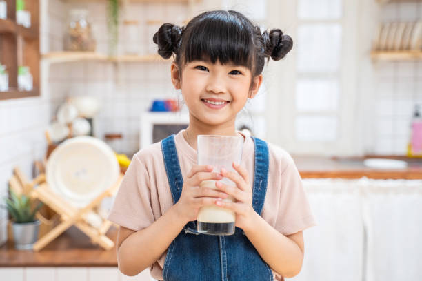 portrait of asian little kid holding a cup of milk in kitchen at home. young preschool cute child girl or daughter stay home with smiling face, feel happy enjoy drinking milk and then look at camera. - milk child drinking little girls imagens e fotografias de stock
