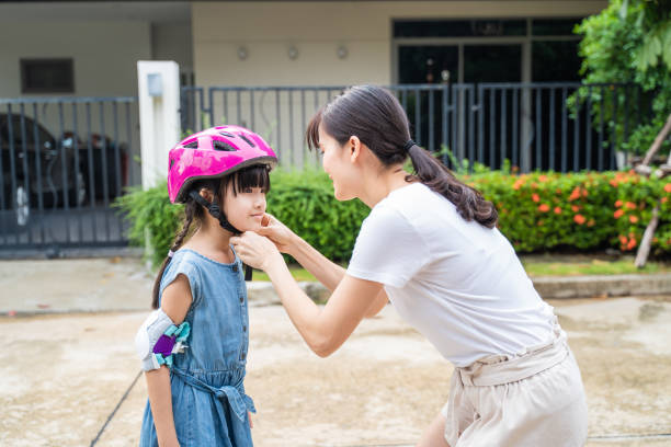 asian mom put on sport helmet to little kid for safety before exercise. beautiful loving mother wear protective hard hat to young girl daughter before ride bicycle for workout on street in the morning. - helmet bicycle little girls child imagens e fotografias de stock