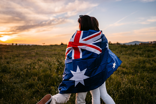 Two, young, females, covering themselves with flag Autralia. Standing on the meadow at sunset. Rear view.