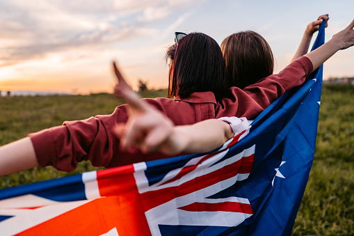 Two, young, females, covering themselves with flag Autralia. Standing on the meadow at sunset. Rear view.