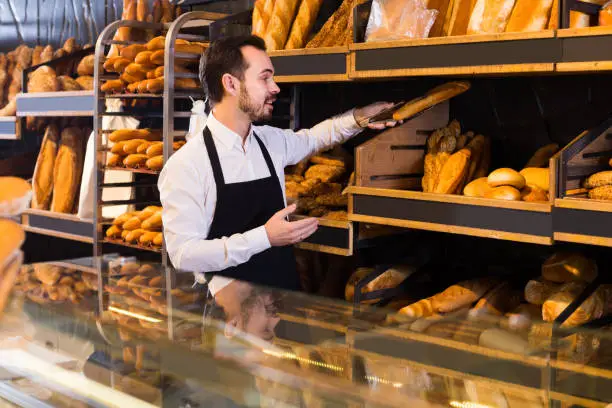 positive spanish male shop assistant demonstrating delicious loaves of bread in bakery