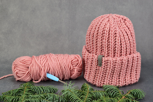 Winter hat, knitted from thick pink yarn. Nearby there are the remains of the yarn, into which the crochet hook is stuck. Image is made on a dark background.