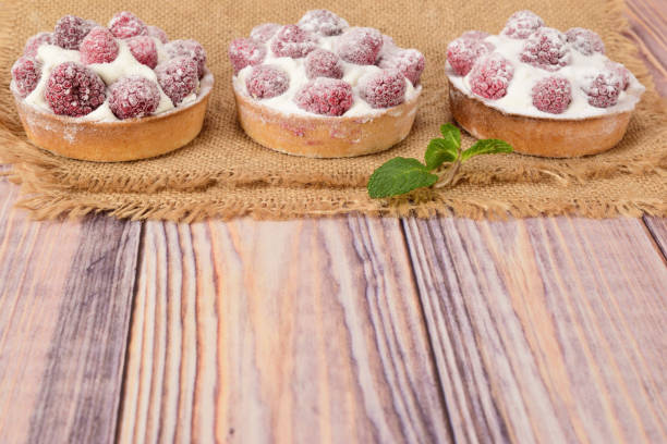 Side view of raspberry tartlets lying on a matting on a wooden table. Cakes sprinkled with powdered sugar, and mint leaves lie next to it.