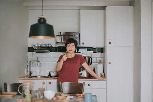 Asian chinese mature woman at kitchen taking a break drinking coffee in the morning for a breakfast and refreshing energy boost before go to work in fresh morning