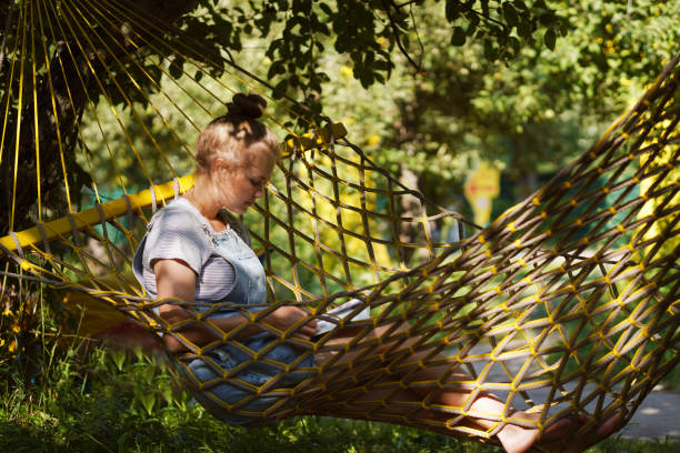 istock Beautiful Young blond woman student smiling Reading A Book While Lying On A Hammock in the public park. Woman relaxing in hammock reading book. Back to school, university, campus concept. 1332091374