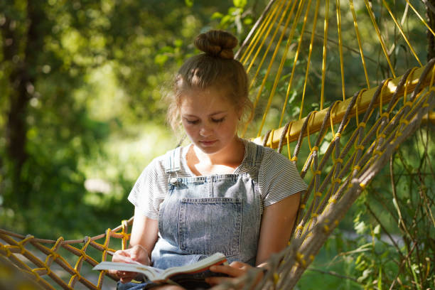 istock Beautiful Young blond woman student smiling Reading A Book While Lying On A Hammock in the public park. Woman relaxing in hammock reading book. Back to school, university, campus concept. 1332091375