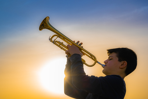 Teen boy blowing Shofar - ram's horn traditionally used for Jewish religious purposes, including the Feast of trumpets, Yom Kippur and Rosh Hashanah; beautiful sunset sky with sunburst in bachground