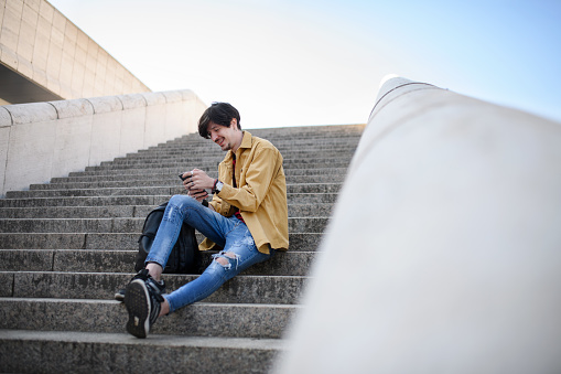 A portrait of young man sitting on staircase outdoors in city, using smartphone