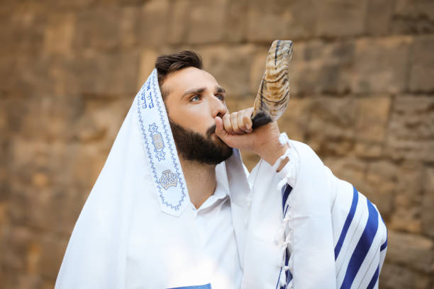 Jewish man blowing shofar on Rosh Hashanah outdoors. Wearing tallit with words Blessed Are You, Lord Our God, King Of The Universe, Who Has Sanctified Us With His Commandments, And Commanded Us To Enwrap Ourselves In Tzitziton in Hebrew hombre judío soplando shofar en rosh hashaná al aire libre. usando tallit con palabras bendito eres tú, señor nuestro dios, rey del universo, que nos ha santificado con sus mandamientos, y nos ha mandado envolvernos en tzitziton en hebreo