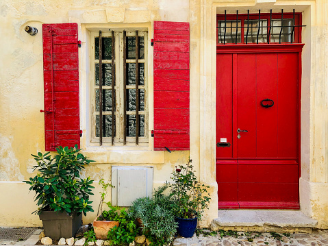 Horizontal closeup photo of an old stone house front facade with a red painted wooden front door and red painted shutters on the window in the Provencal city of Arles on a sunny day in Spring