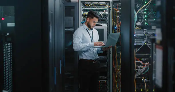Photo of Shot of a young male engineer using his laptop in a server room