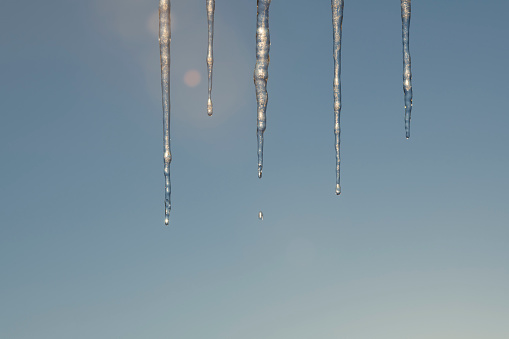 Backlit icicles hanging from rooftop are melting in the afternoon sun in winter (or early spring). The image contains lens flare (that adds to the atmosphere).