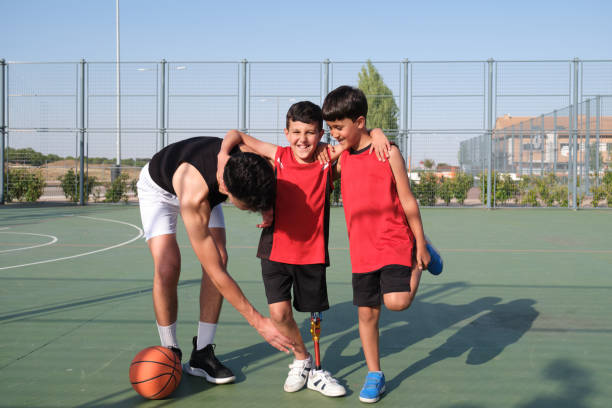 Two brothers helping to their sibling with a leg prosthesis to stretch his leg. Two brothers helping to their sibling with a leg prosthesis to stretch his other leg. Siblings stretching before playing basketball. athlete with disabilities photos stock pictures, royalty-free photos & images