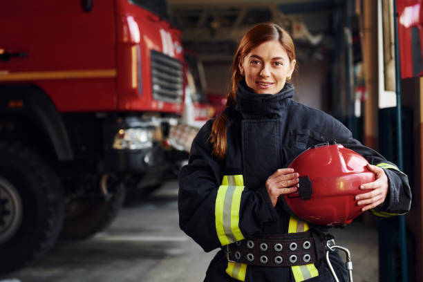 Holds red hat in hands. Female firefighter in protective uniform standing near truck stock photo