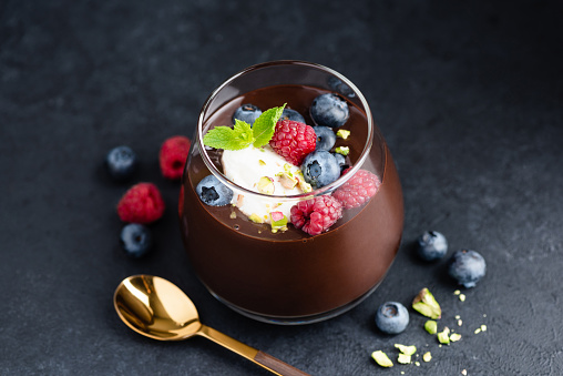 Dark chocolate mousse with pistachios, berries and cream in jar, black stone background, closeup view