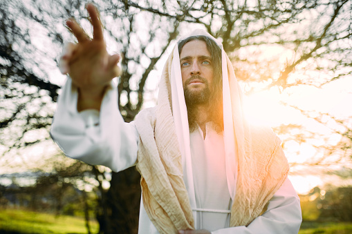 Jesus Christ standing in meadow and preaching Christian faith conversion clothed in white robe against tree and sunset background. Portrait.