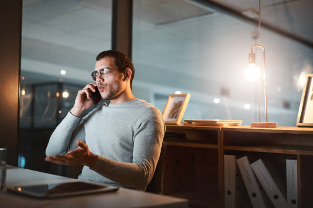 istock Shot of a young man talking on his cellphone while using his computer at night at the office 1332138703