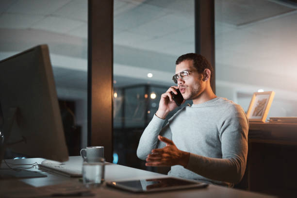 istock Shot of a young man talking on his cellphone while using his computer at night at the office 1332138707