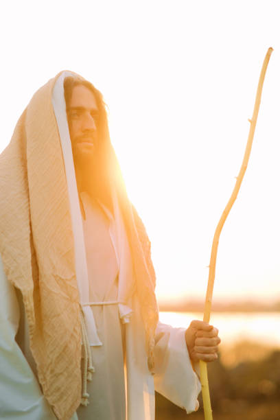 istock Jesus Christ with staff stands in white robe near river at sunset. 1332138708