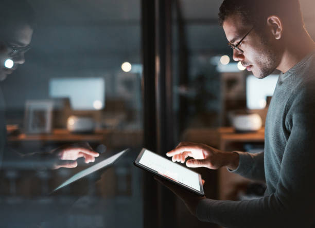 istock Shot of a young man standing at his office window and using his digital tablet at night 1332139242