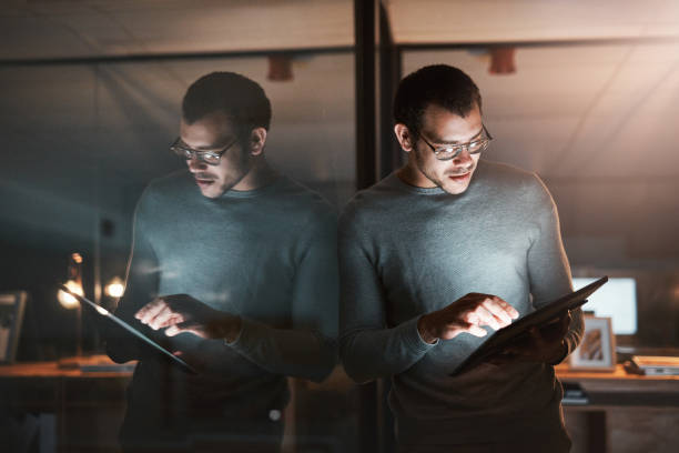istock Shot of a young man standing at his office window and using his digital tablet at night 1332139245