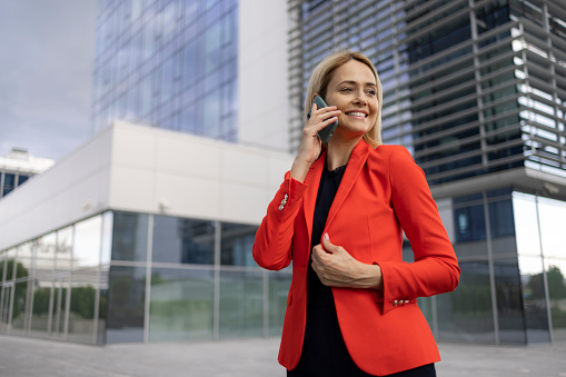 Happy middle aged Caucasian businesswoman wearing a red female business suit smiling and walking in front of the office building, using a smart phone
