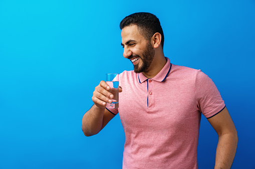Handsome hispanic man drinking a glass of fresh water against blue background, close up