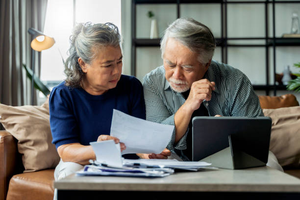 vecchia coppia asiatica senior in pensione che controlla e calcola la fatturazione finanziaria insieme sul divano coinvolto in scartoffie finanziarie, pagando le tasse online utilizzando il laptop e-banking sul background della casa del soggiorno - retirement senior adult planning finance foto e immagini stock