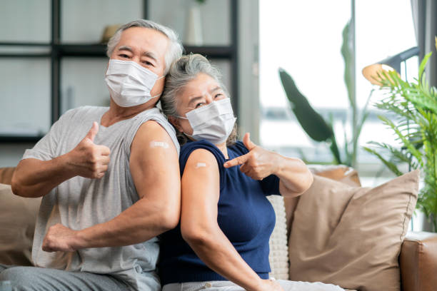 old asian senior couple wearing face mask virus protection happiness cheerful show bandage injection covid-19 vaccinated with hand gesture point and confident for virus protection stock photo