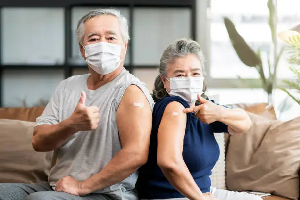 Photo of old asian senior couple wearing face mask virus protection happiness cheerful show bandage injection covid-19 vaccinated with hand gesture point and confident for virus protection