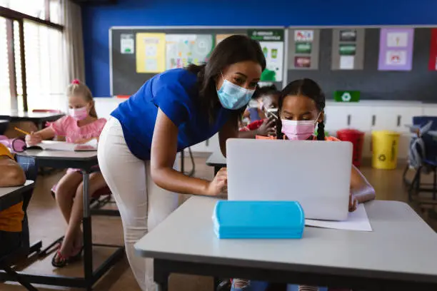 African american female teacher wearing face mask teaching a girl to use laptop at elementary school. back to school and education concept