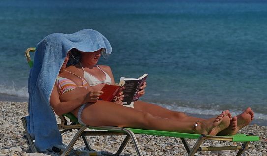 Samos Island, Greece - August 18, 2006: Two girls sharing the same sunbed and use the same towel for some shade, are reading their books on a hot August day at the crowded Potokaki beach.