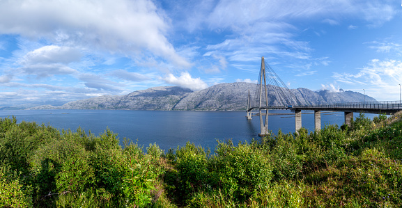 Panoramic view at Helgeland Bridge over blue wide river and Seven Sisters mountain range in summer sunny day under blu sky. Norway's finest bridges between Alstahaug and Leirfjord at Helgeland, Norway. At Sandnessjoen town.