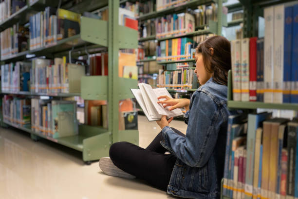 istock Asian female student sitting on floor in the library, Open and learning textbook from bookshelf 1332169111