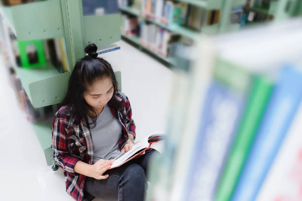 istock Asian female student sitting on floor in the library, Open and learning textbook from bookshelf 1332169126