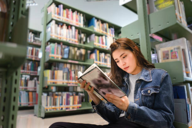 istock Asian female student sitting on floor in the library, Open and learning textbook from bookshelf 1332169127