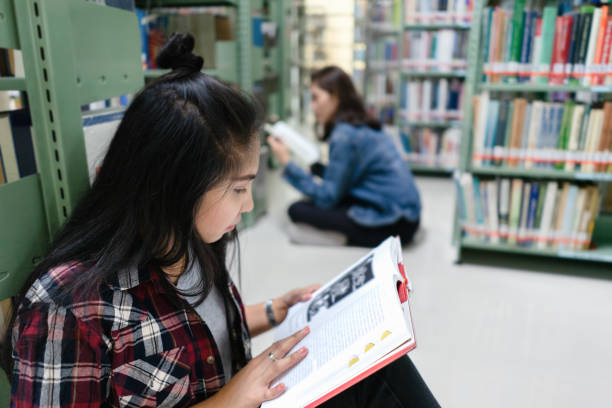 istock Asian female student sitting on floor in the library, Open and learning textbook from bookshelf 1332169130