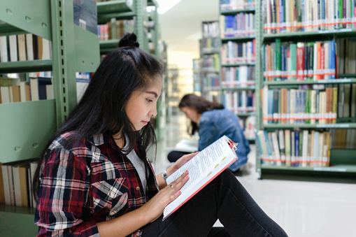 Asian female student sitting on floor in the library, Open and learning textbook from bookshelf in the International College/University Library.