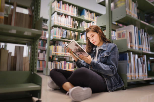 asian female student sitting on floor in the library, open and learning textbook from bookshelf - library stockfoto's en -beelden